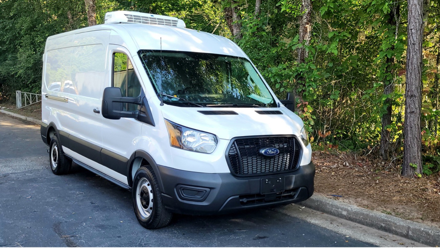 White Ford Transit refrigerated van with rooftop cooling system, parked near trees.